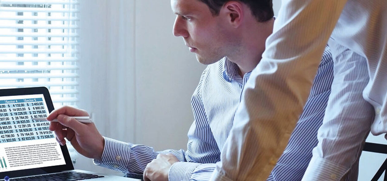 Finanzbuchhalter beim Austausch mit einer anderen Person vor einem Laptop im Büro.