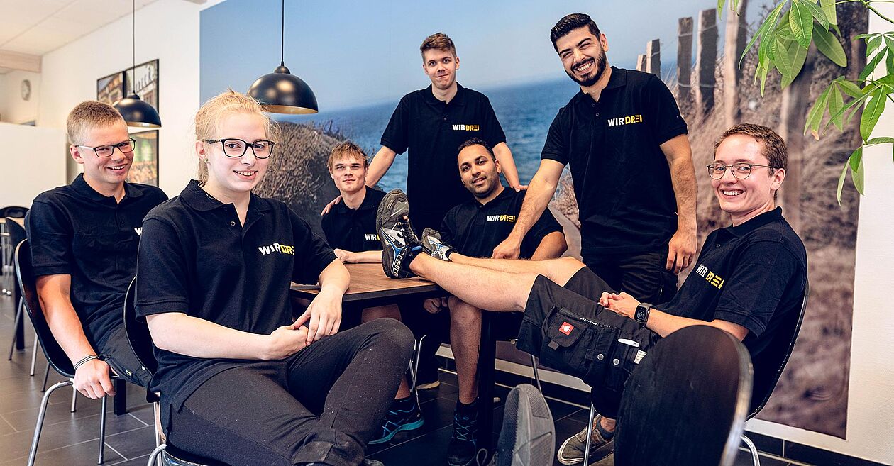 Teamfoto der WIR DREI Mitarbeiter an einem Tisch mit Stühlen.