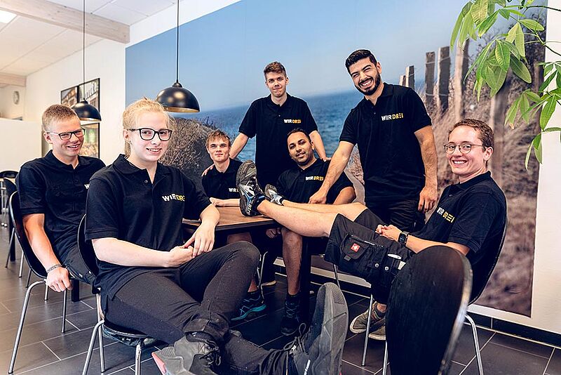 Teamfoto der WIR DREI Mitarbeiter an einem Tisch mit Stühlen.