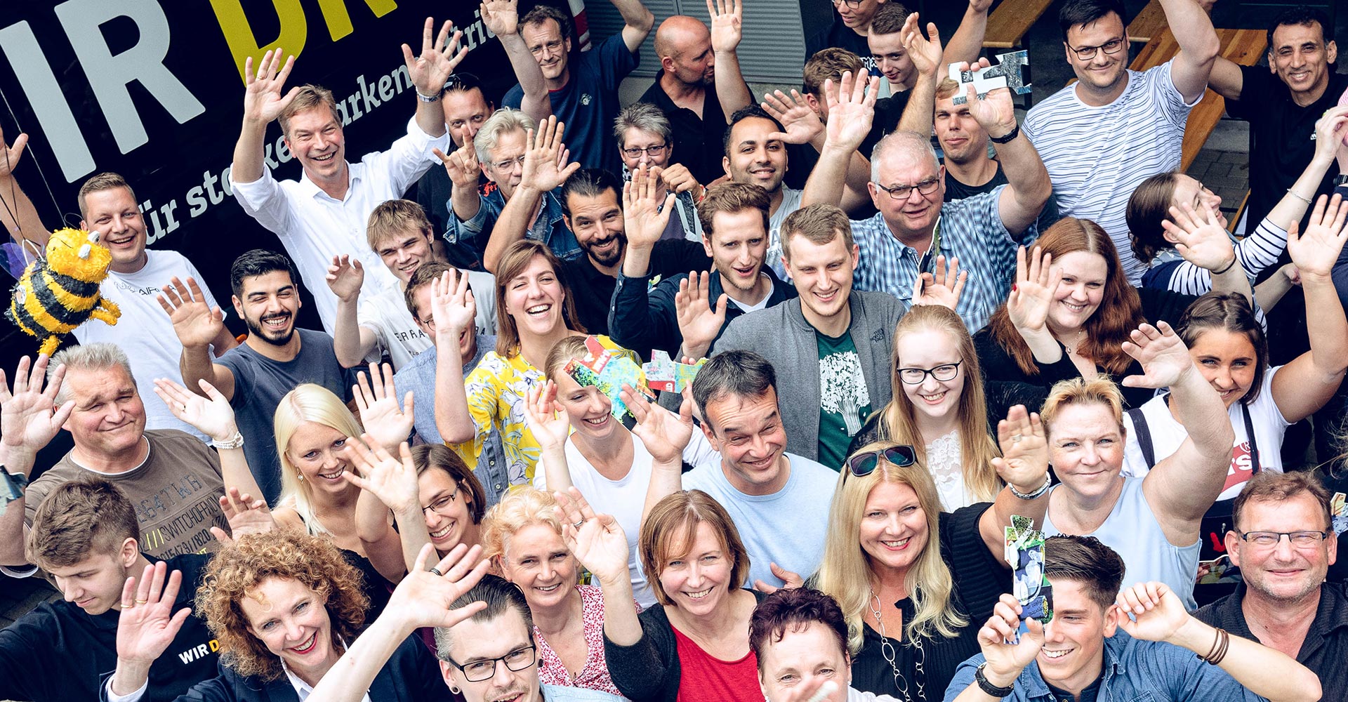 Das WIR DREI Team von oben während alle mindestens eine Hand heben und in die Kamera lächeln.